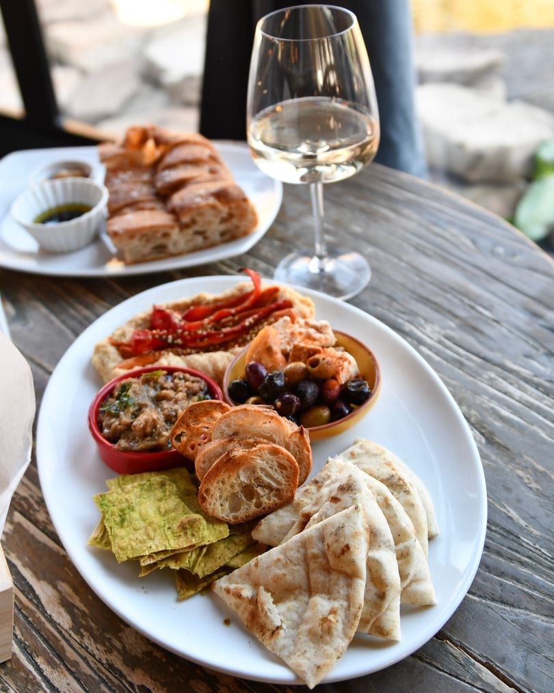 Appetizer platter with various dips, olives, pita bread, and baguette slices, with a glass of white wine.