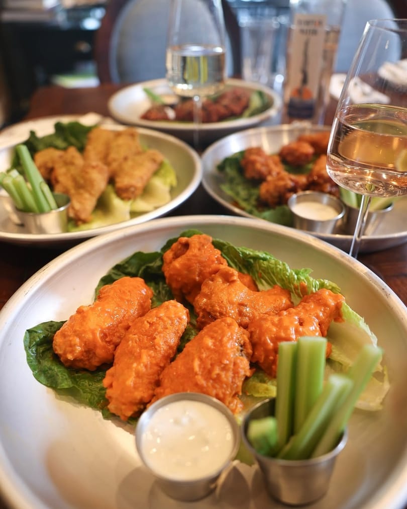 Plates of buffalo wings with celery sticks and dipping sauce, with wine glasses on a table.