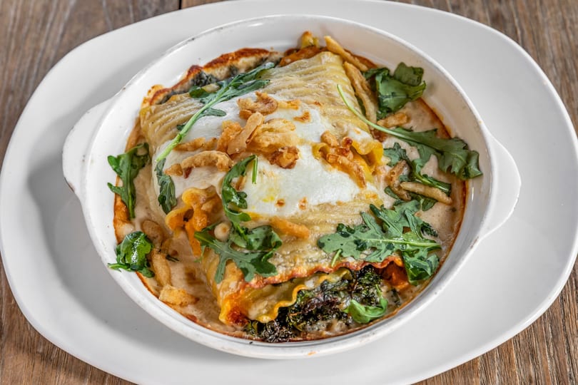 Baked lasagna with melted cheese, arugula, and crispy fried onions in a white dish.