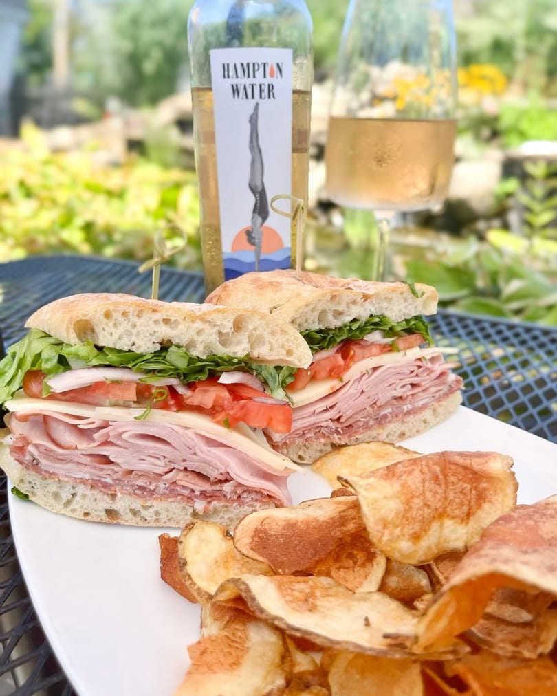 Deli sandwich with chips, a bottle of Hampton Water rosé, and a glass of wine outdoors.